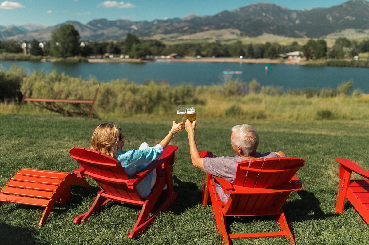 A couple cheers-ing on MAP Brewing Company's lawn overlooking a lake and the Bridger Mountains.