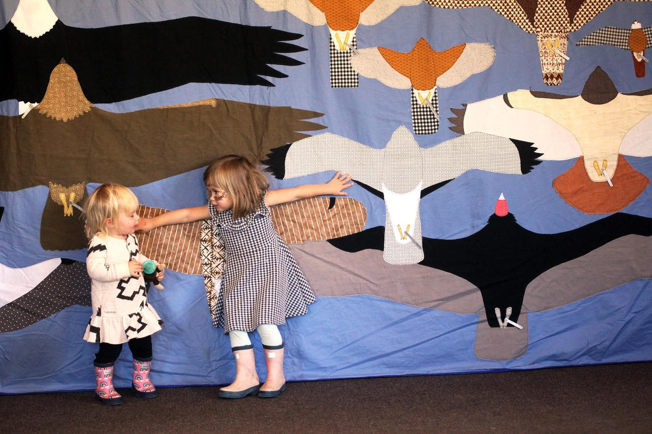 two children compare their wingspans to those of birds on a quilt behind them